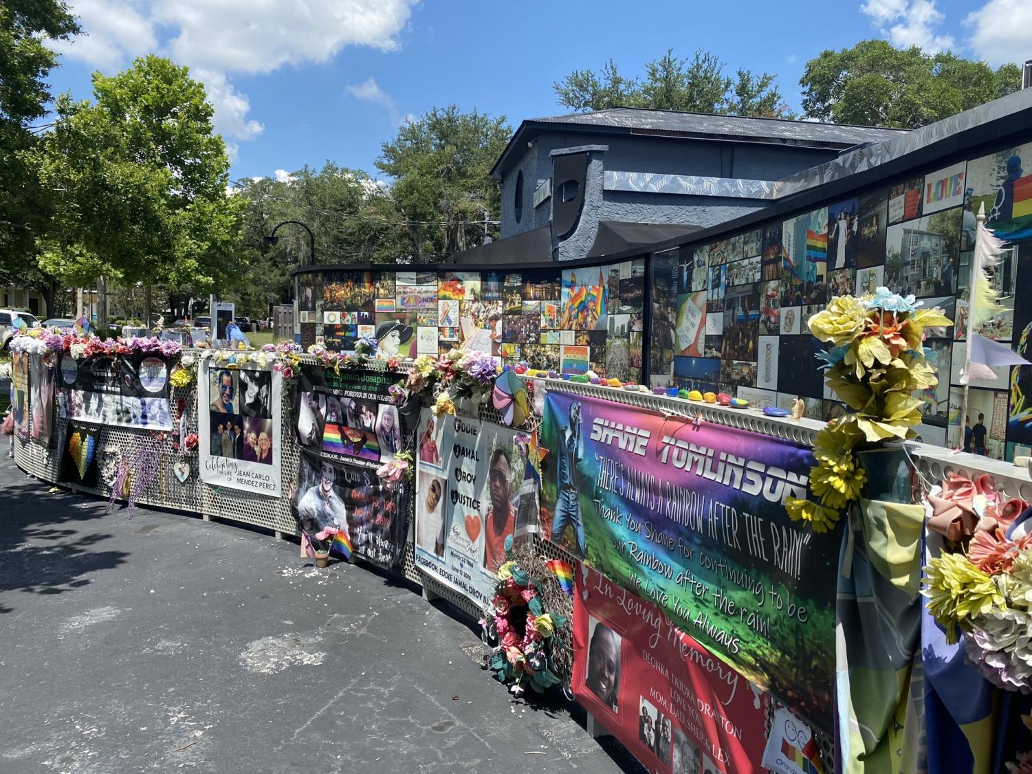 The Interim Pulse Memorial Wall extends along the outside of the Pulse nightclub exterior with window panes for looking inward. Names of the 49 lives lost are visible on the wall inside. 